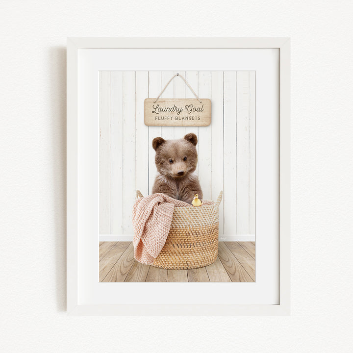 A framed image of a brown bear sitting in a woven basket, with a sign above the basket that reads "Laundry Goal Fluffy Blankets".