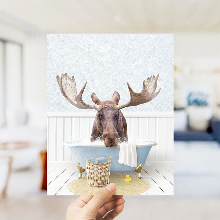 A moose with large antlers is sitting in a bathtub filled with water, surrounded by a yellow rubber duck and a wicker basket.