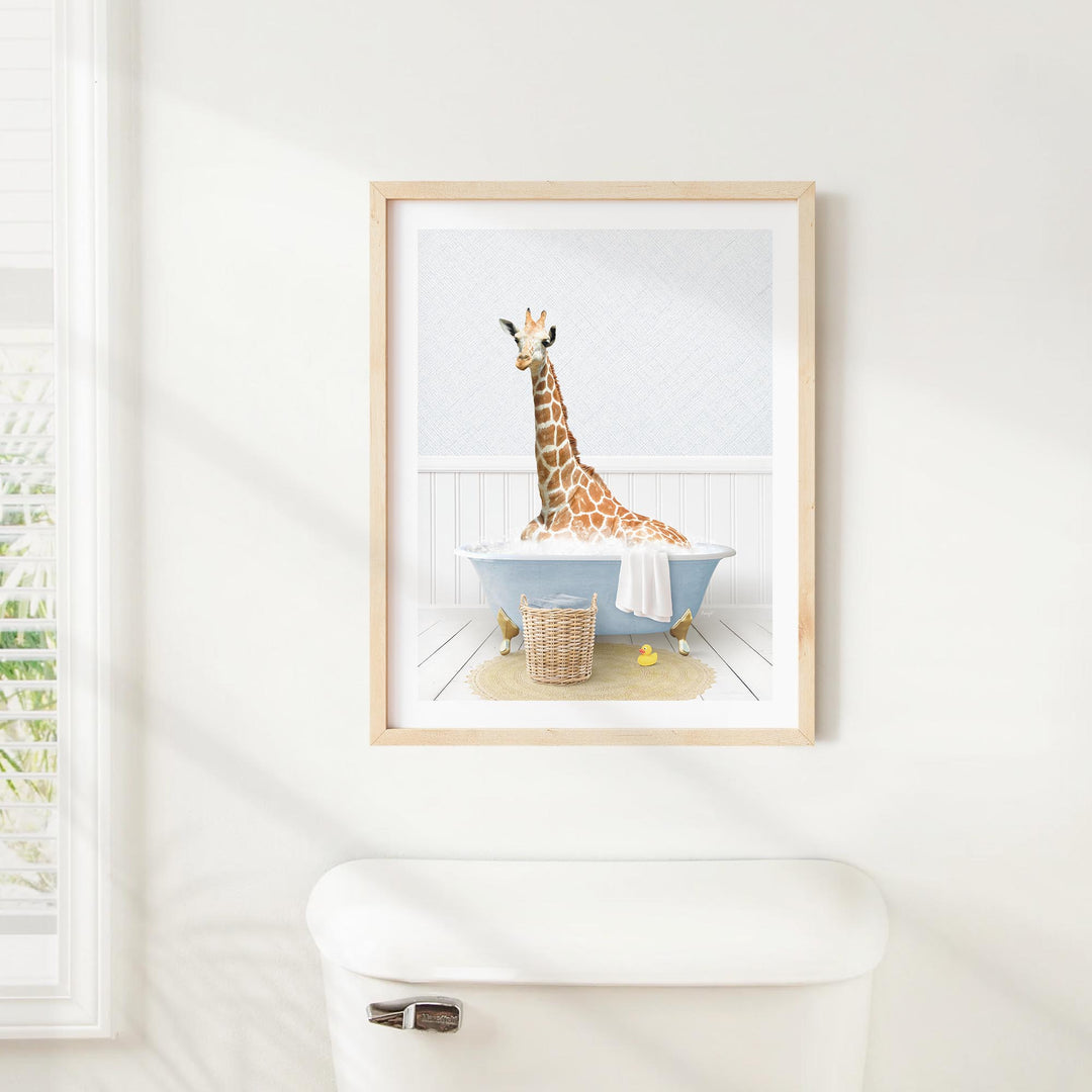 A giraffe sitting in a blue bathtub, surrounded by a basket of toys and a towel, is the central focus of the framed image displayed on the wall.