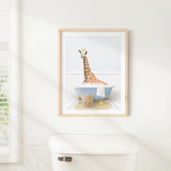 A giraffe sitting in a blue bathtub, surrounded by a basket of toys and a towel, is the central focus of the framed image displayed on the wall.