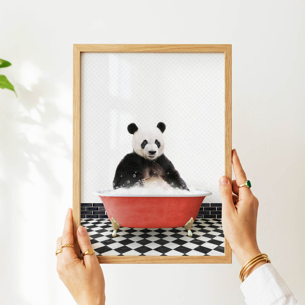 A person's hands are holding a framed picture of a panda bear sitting in a red bathtub filled with bubbles.