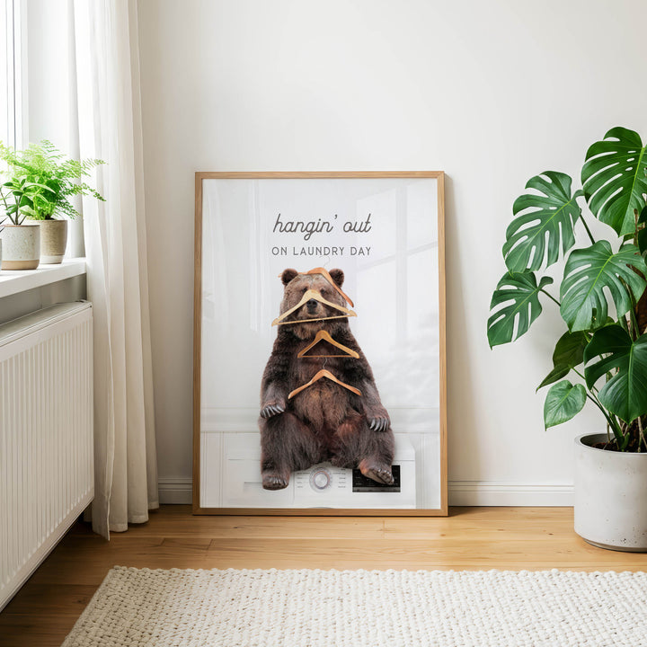A framed poster with a bear sitting on a washing machine, surrounded by clothes hangers, is displayed in a room with a window and potted plants.