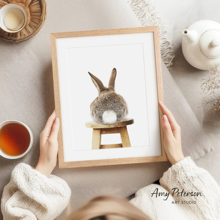 A person is holding a framed picture of a rabbit sitting on a wooden stool, with a teapot and a cup of tea nearby.