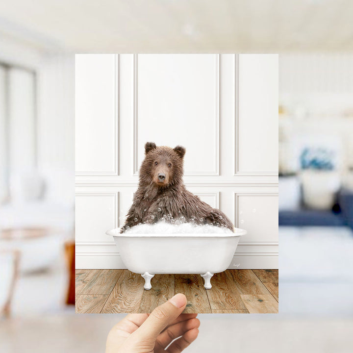 A brown bear sitting in a white bathtub filled with bubbles, with a blurred background suggesting a bathroom setting.