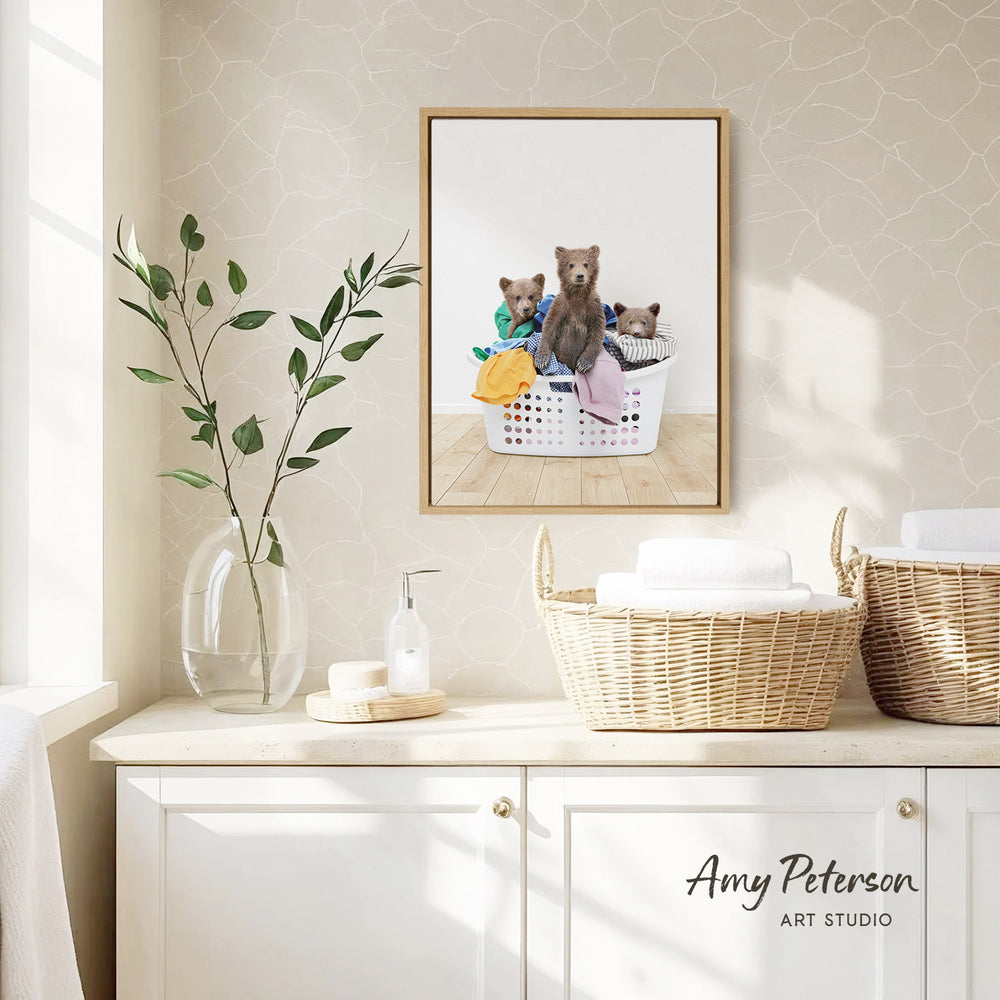 a framed photograph of three bears sitting in a laundry basket, with a vase of leaves and a bottle of soap on a white cabinet.