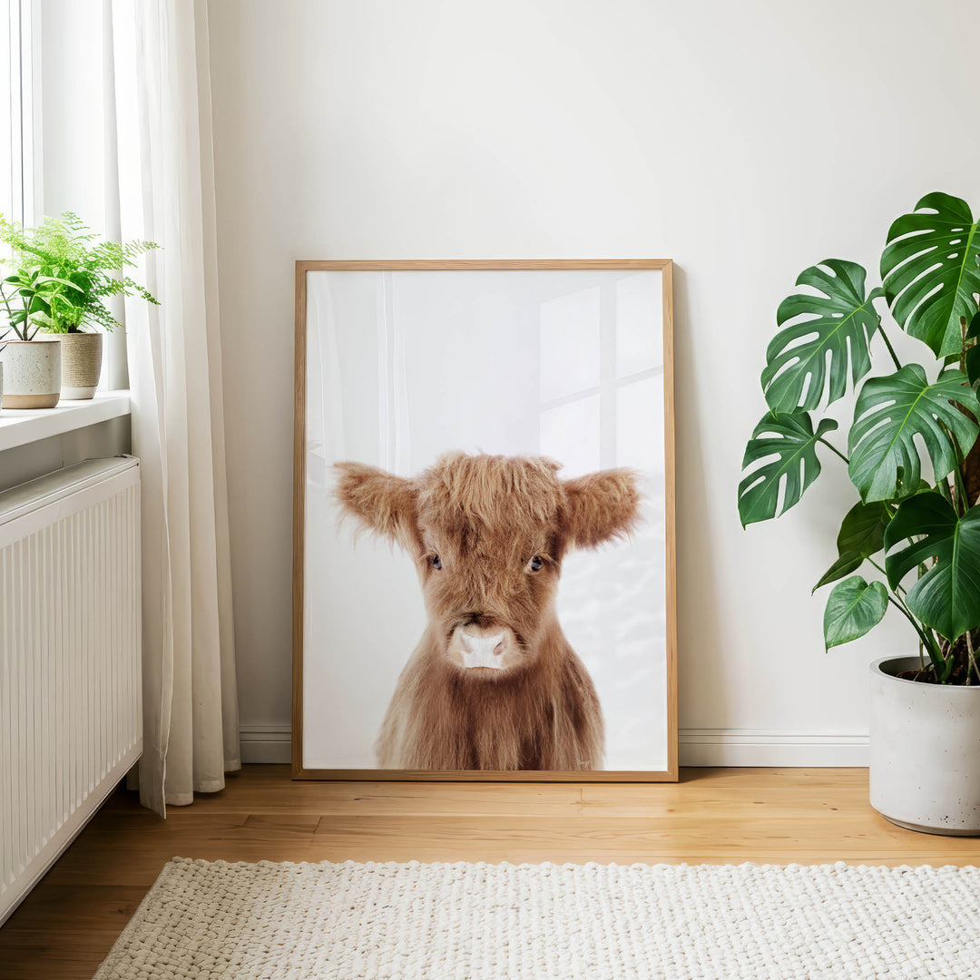 a framed picture of a brown cow with large ears, placed on a wooden floor in a room with a window and potted plants.