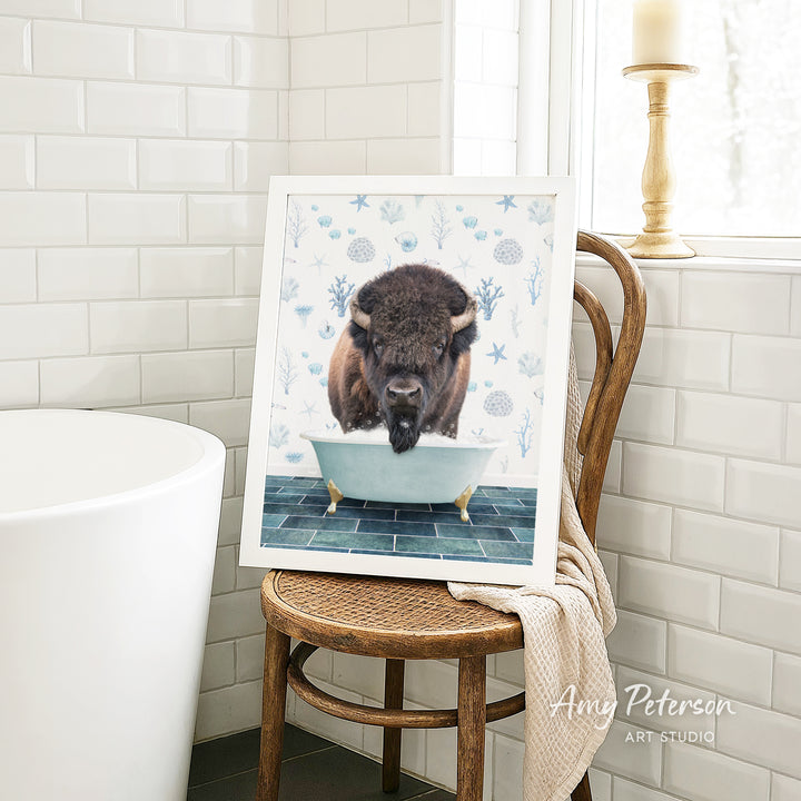 A framed image of a buffalo standing in a bathtub, with a towel draped over the back of a wooden chair in a bathroom setting.