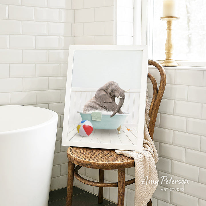 A framed image of a gray elephant sitting in a blue bathtub, surrounded by a beach ball, a towel, and a candle, is displayed on a wooden chair in a bathroom setting.