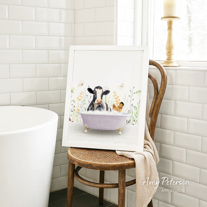 A framed painting depicting a cow and a chicken relaxing in a bathtub, surrounded by flowers, is displayed on a wooden chair in a bathroom setting.
