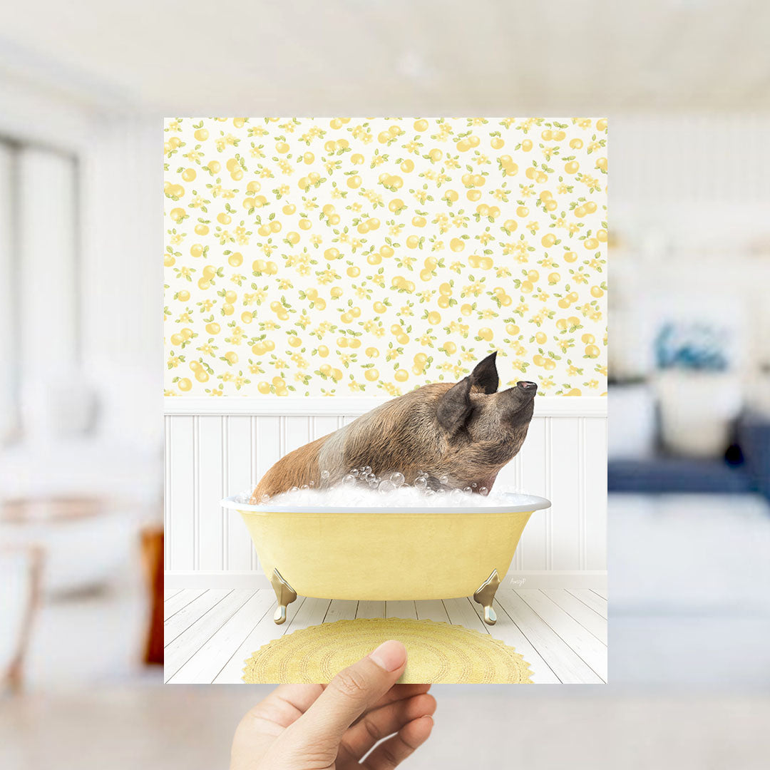 A hand holds up a framed image depicting a pig sitting in a yellow bathtub filled with bubbles, surrounded by a yellow and white floral pattern.