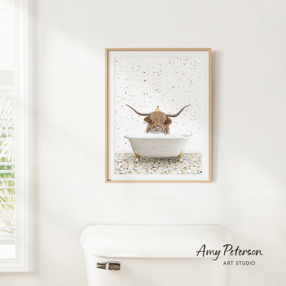 A framed print of a bull with horns sitting in a bathtub, surrounded by a speckled background, hangs on a white wall.