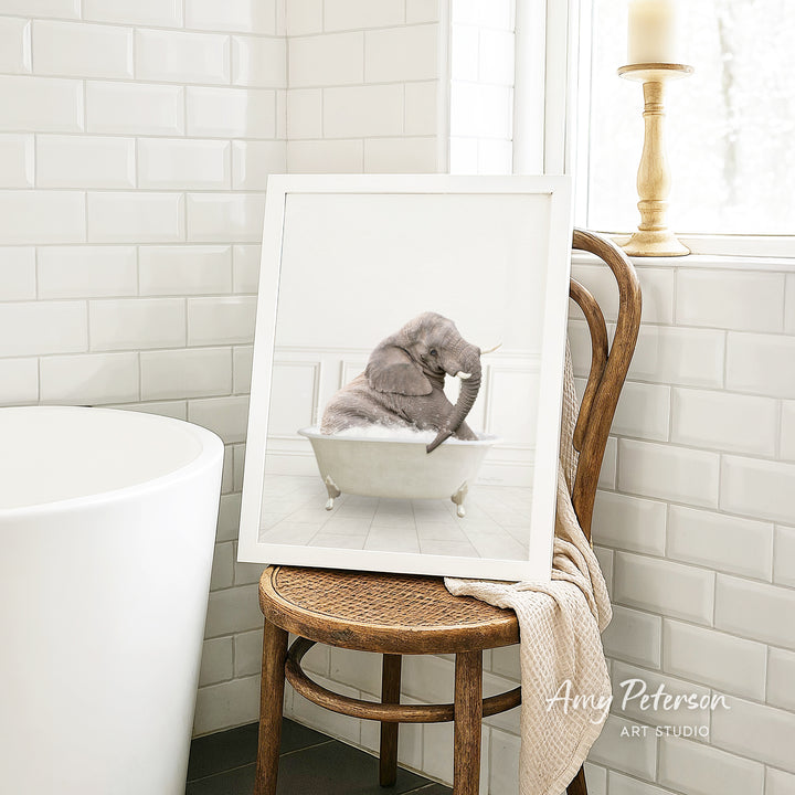 A framed image of a cat sitting in a bathtub, with a towel draped over the back of a wooden chair in a bathroom setting.
