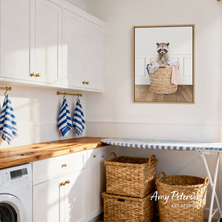 A laundry room with white cabinets, a wooden countertop, and a framed picture of a raccoon in a basket. There are also two woven baskets and a striped ironing board in the room.