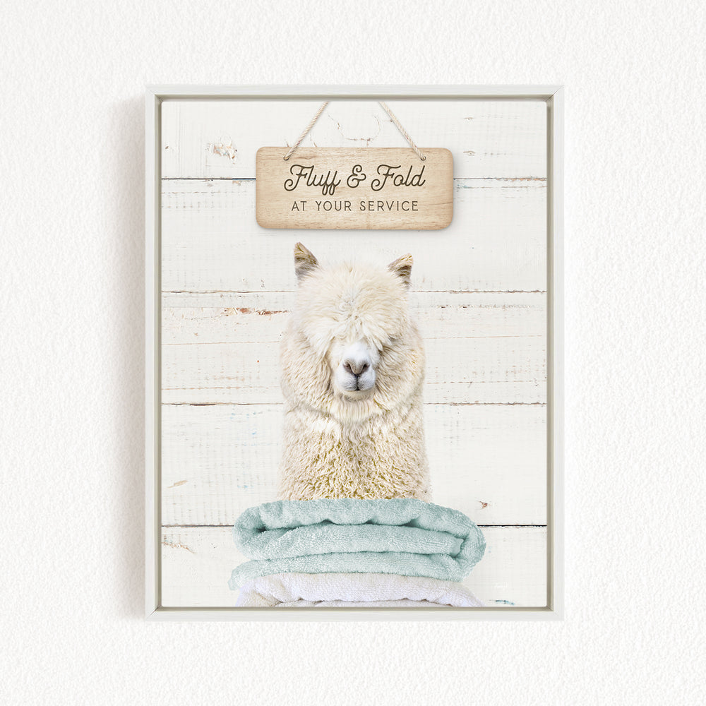 A framed image of a fluffy white alpaca sitting on a stack of light blue towels, with a wooden sign above it that says "Fluff & Fold at your service".