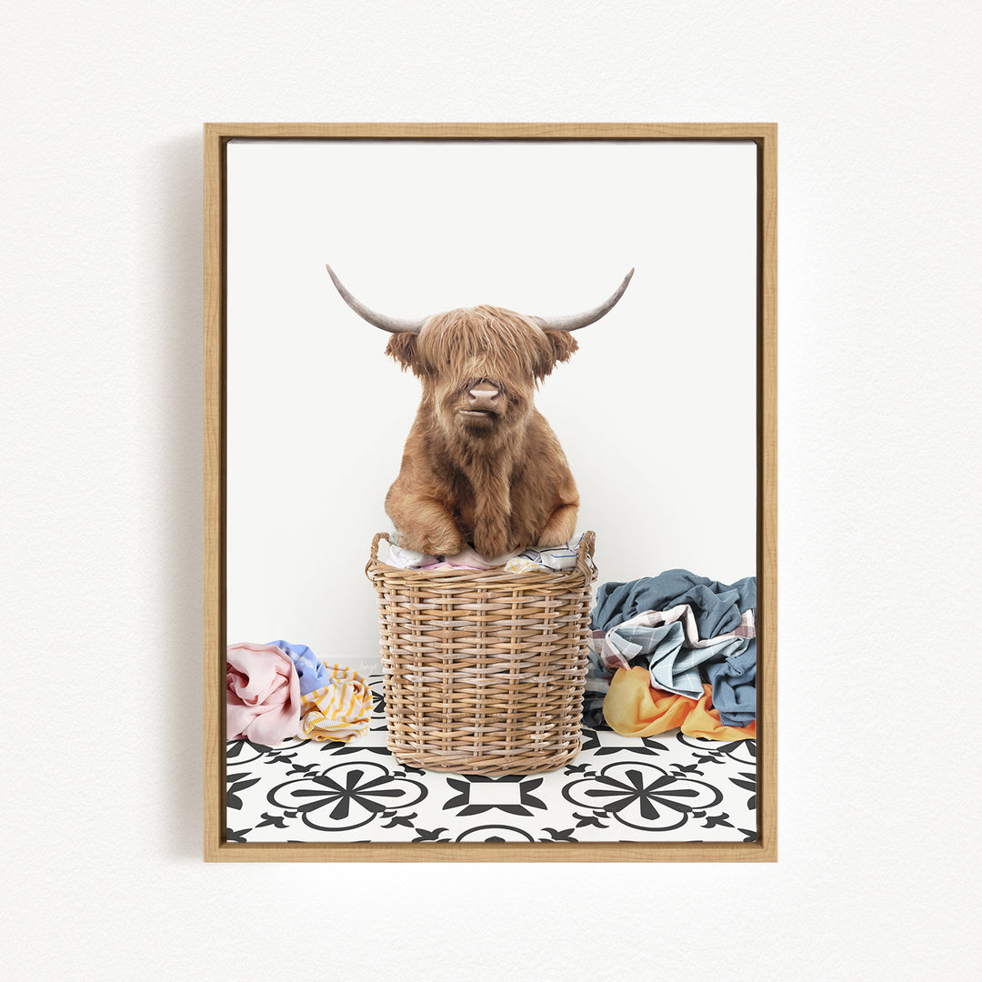 A Highland cow with long horns is sitting in a wicker basket filled with clothes, placed on a patterned floor.