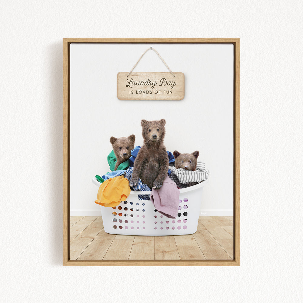 A wooden sign with the text "Laundry Day is loads of fun" hangs above a white laundry basket filled with clothes, where three bears are sitting and resting.