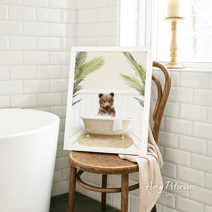 A framed image of a teddy bear sitting in a bathtub, surrounded by palm leaves, is displayed on a wooden chair in a bathroom setting.