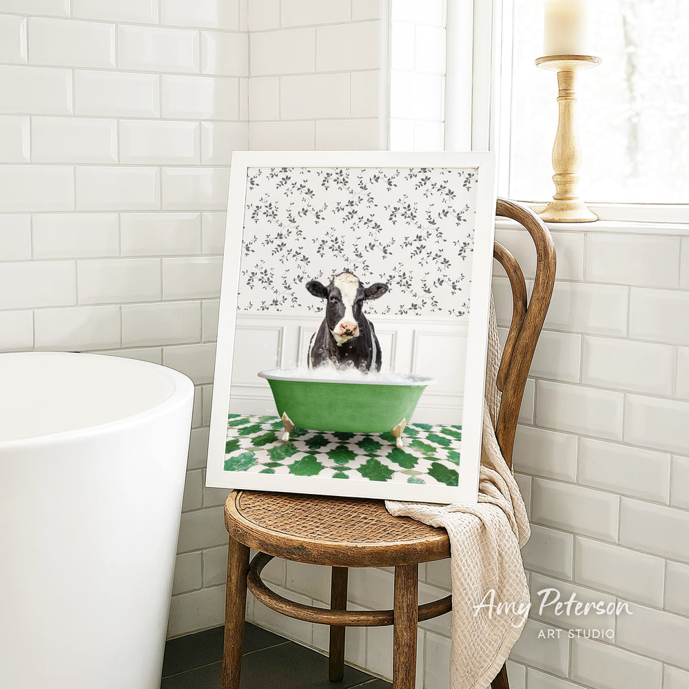 A framed image of a cow sitting in a green bathtub, surrounded by a floral pattern, is displayed on a wooden chair in a bathroom setting.