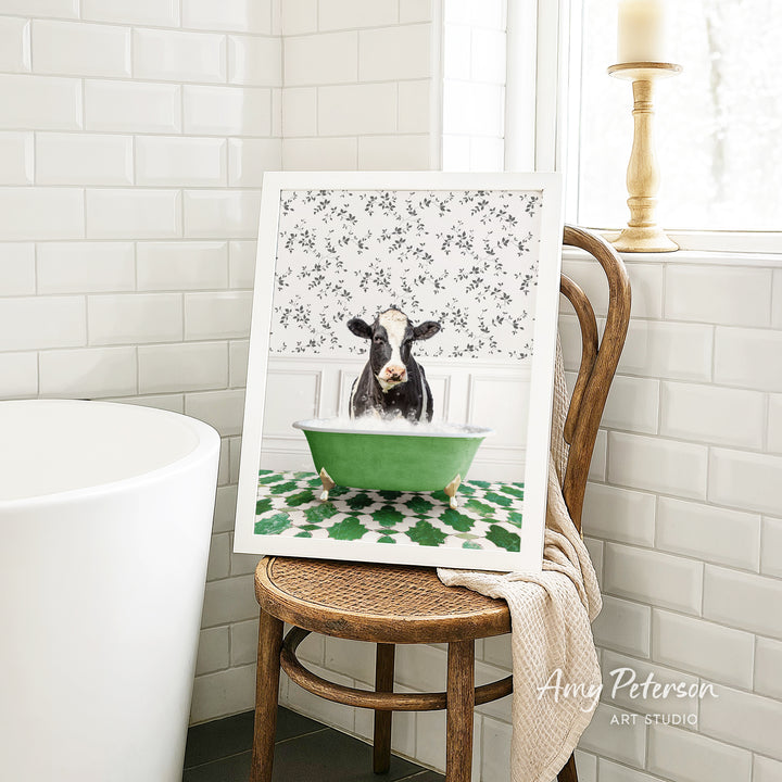 A framed image of a cow sitting in a green bathtub, surrounded by a floral pattern, is displayed on a wooden chair in a bathroom setting.