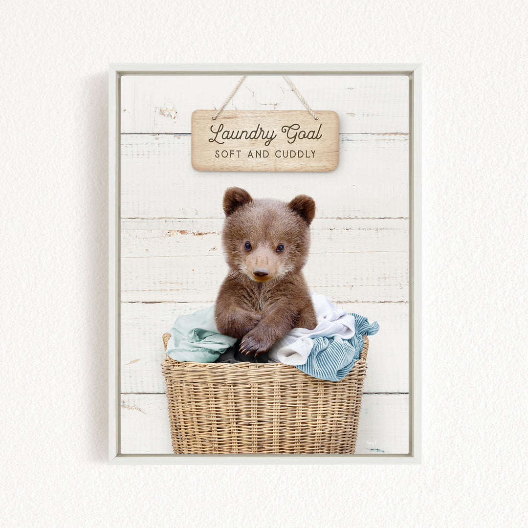 A cute brown bear sitting in a wicker laundry basket, with a wooden sign above that says "Laundry Goal Soft and Cuddly".