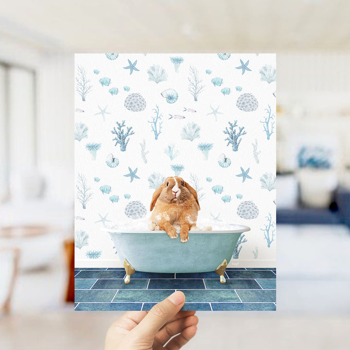 A hand is holding up a print featuring a brown rabbit sitting in a bathtub filled with bubbles, surrounded by a pattern of seashells, starfish, and other marine life.