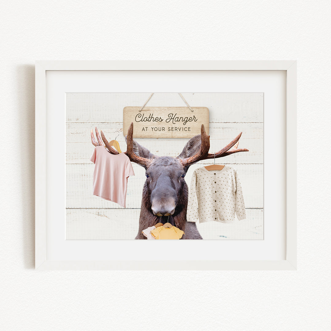 A framed image depicting a moose with antlers holding clothes, including a pink shirt and a white sweater, against a white background with a sign that reads "Clothes Hanger at your service".