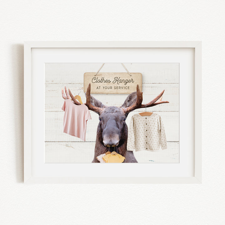 A framed image depicting a moose with antlers holding clothes, including a pink shirt and a white sweater, against a white background with a sign that reads "Clothes Hanger at your service".