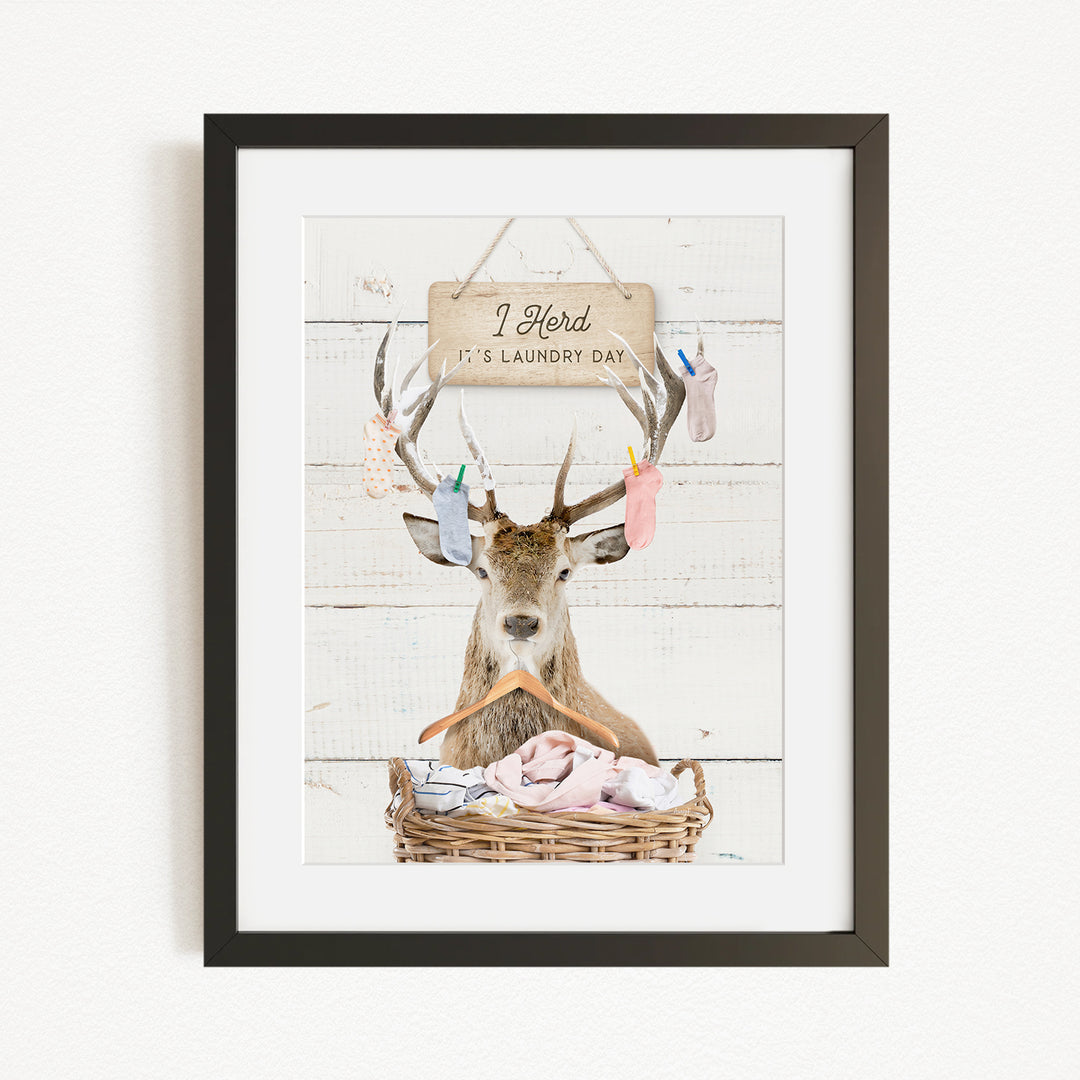 A framed image depicting a deer with antlers holding laundry, accompanied by a sign that reads "I herd it's laundry day".