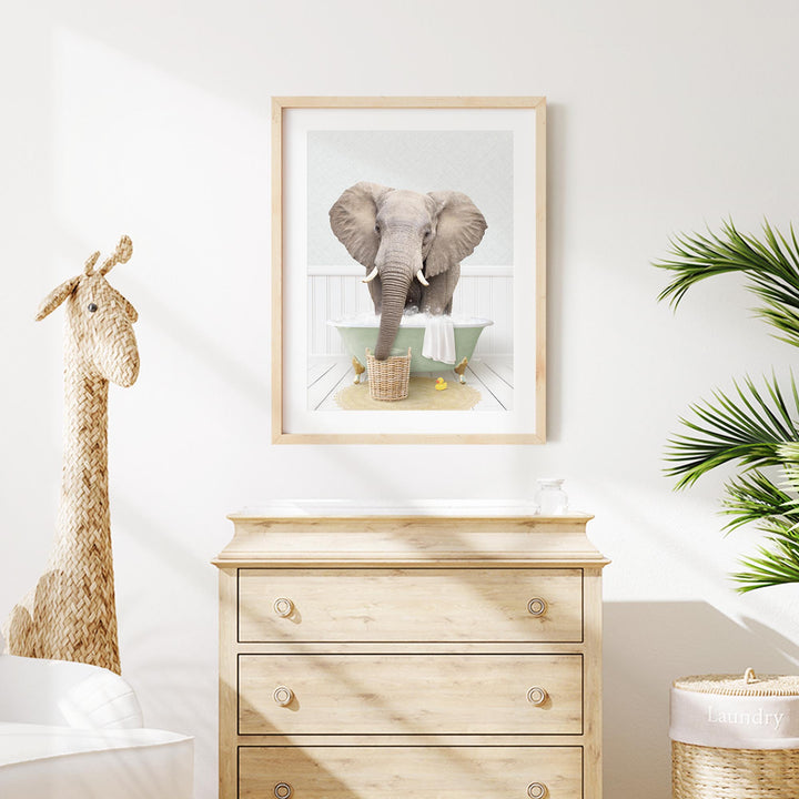 a framed photograph of an elephant standing in a bathtub, with a giraffe figurine next to it. The photograph is hanging on a white wall above a wooden dresser.