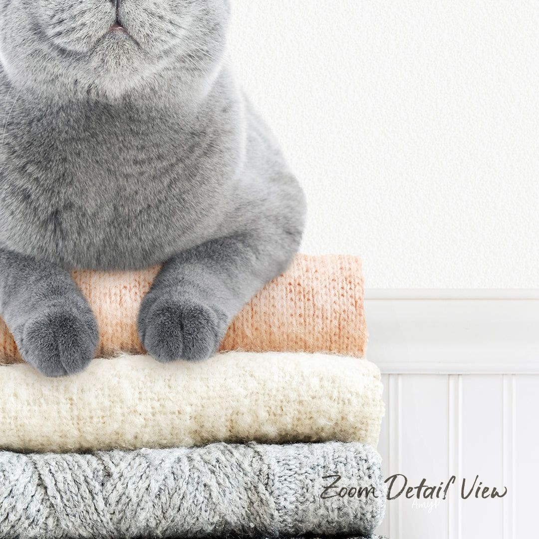 A gray cat sitting on top of a stack of three different colored sweaters.