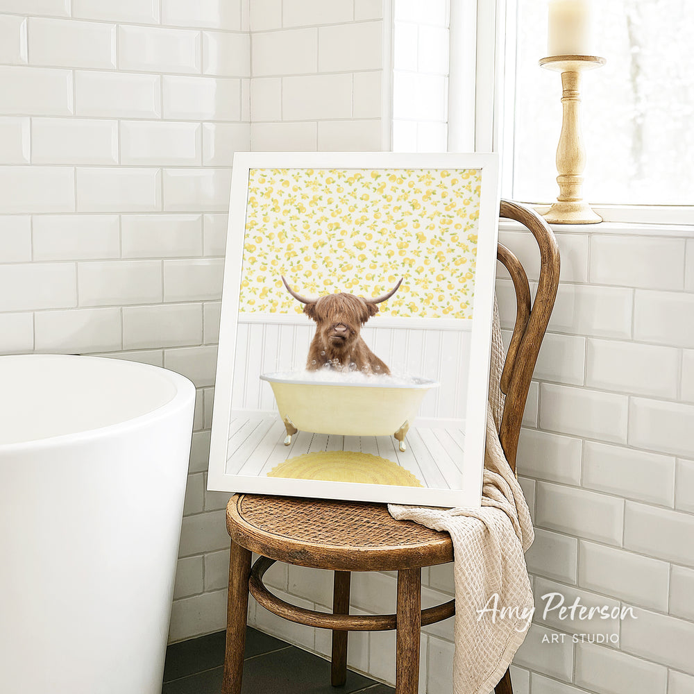 A framed image of a bull with horns sitting in a bathtub, surrounded by yellow flowers, is displayed on a wooden chair in a bathroom setting.