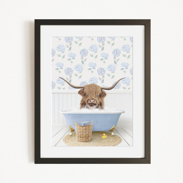 A framed image depicting a Highland cow with long horns sitting in a blue bathtub, surrounded by a floral patterned wall and a basket of rubber ducks.