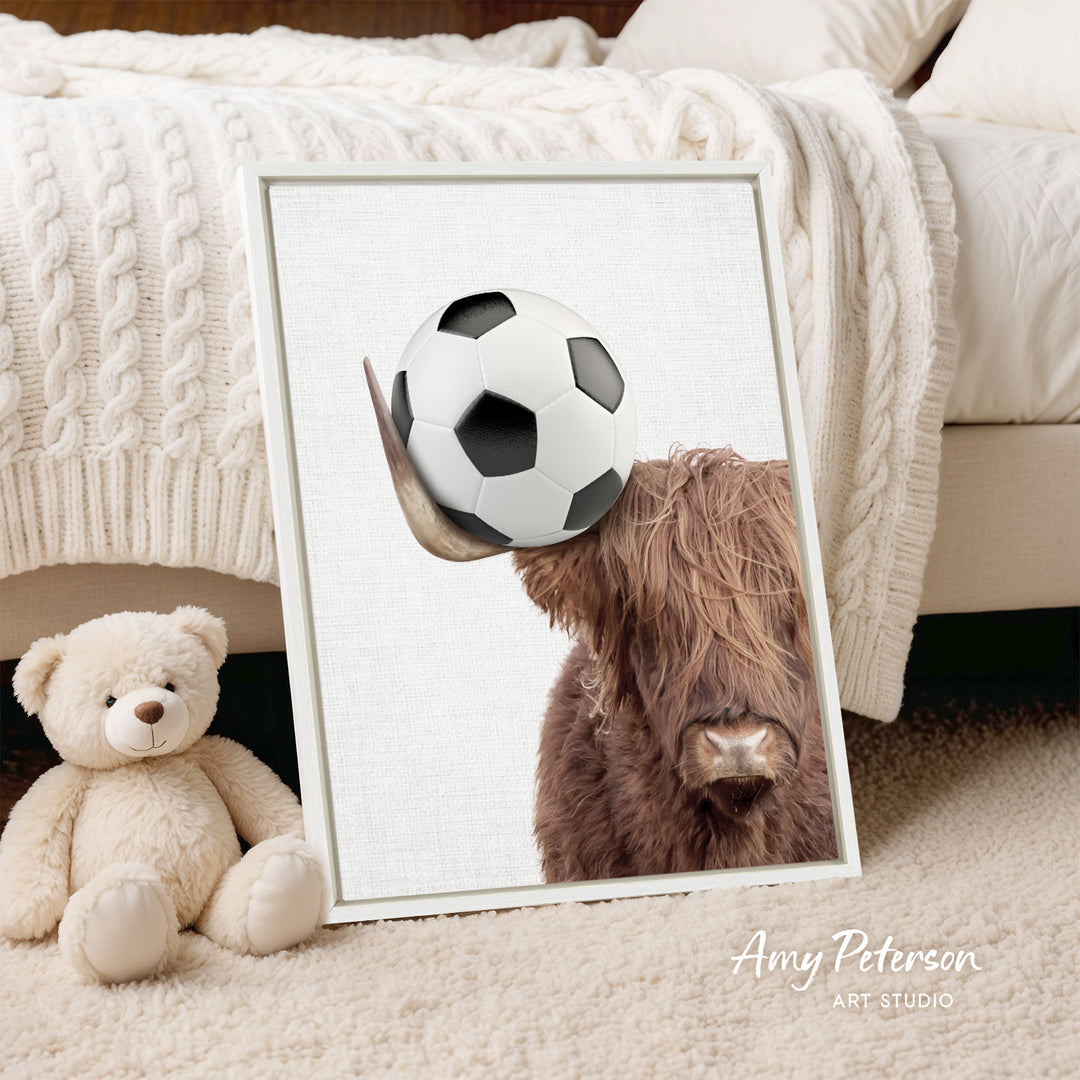 A framed image of a Highland cow with a soccer ball in its mouth, placed on a bed next to a teddy bear.
