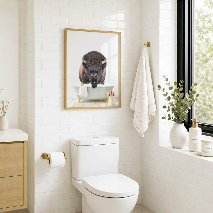 A bathroom with a white toilet, a framed picture of a buffalo in a bathtub, and a window with greenery outside.