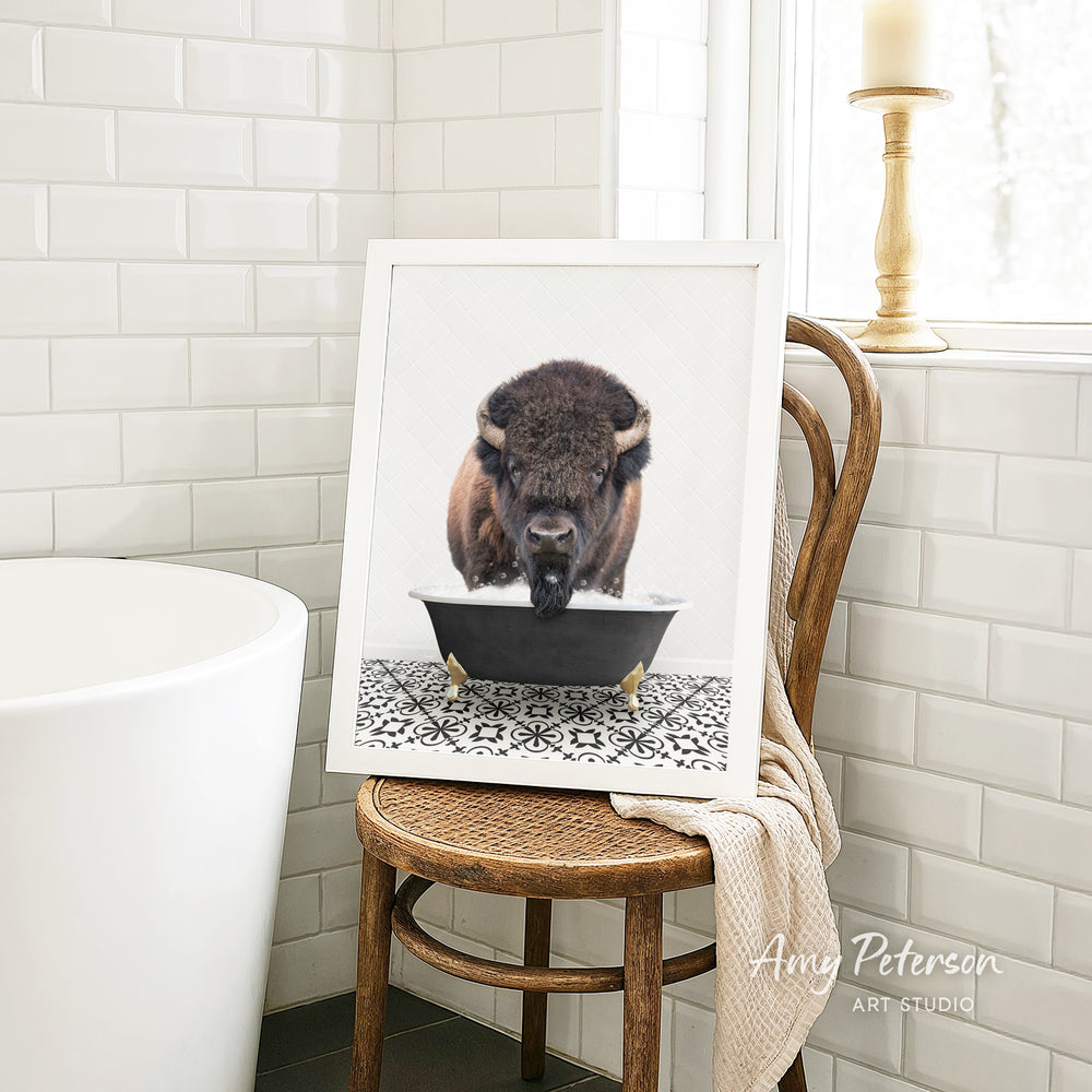 A framed image of a bison resting in a bathtub is displayed on a wooden chair in a bathroom setting.