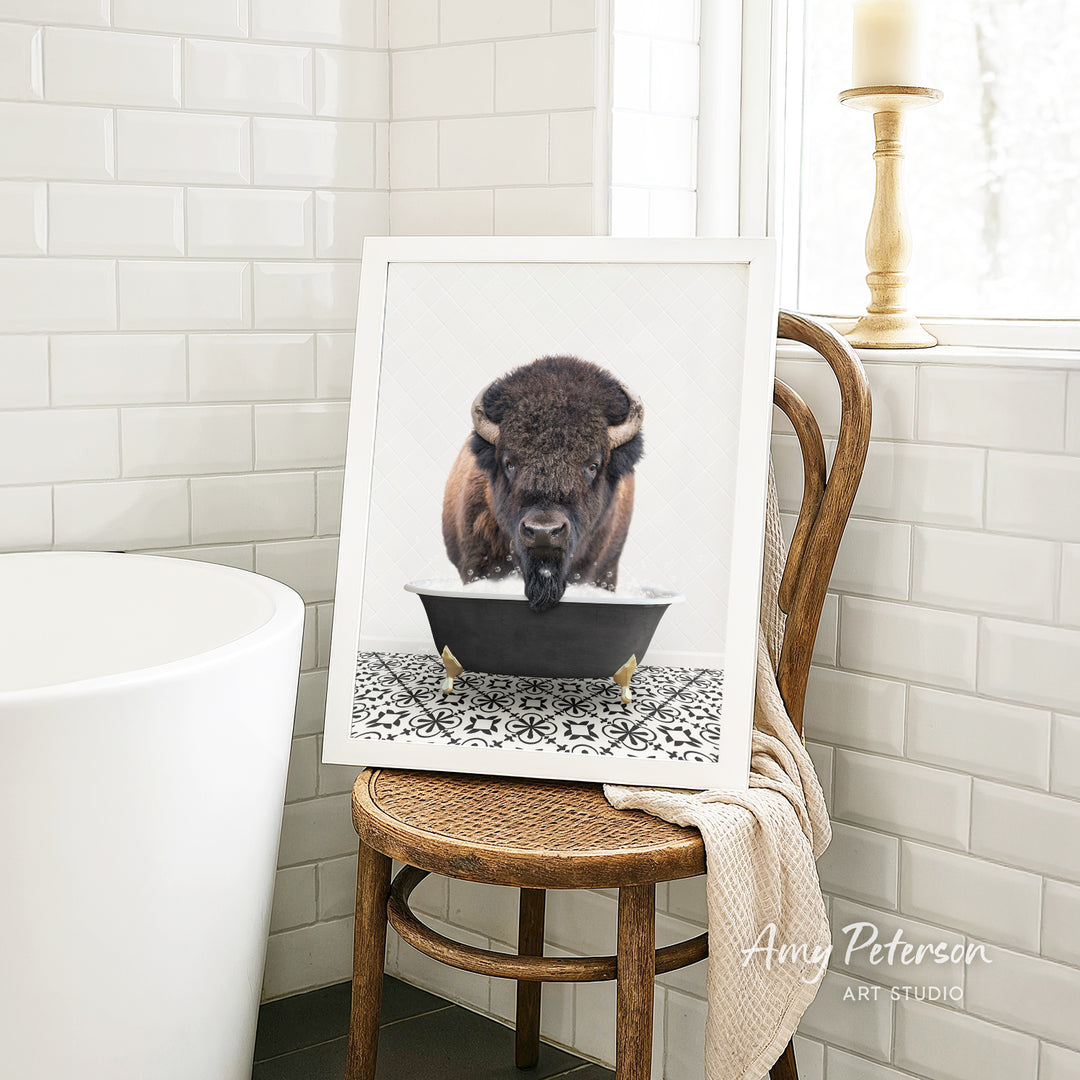 A framed image of a bison resting in a bathtub is displayed on a wooden chair in a bathroom setting.