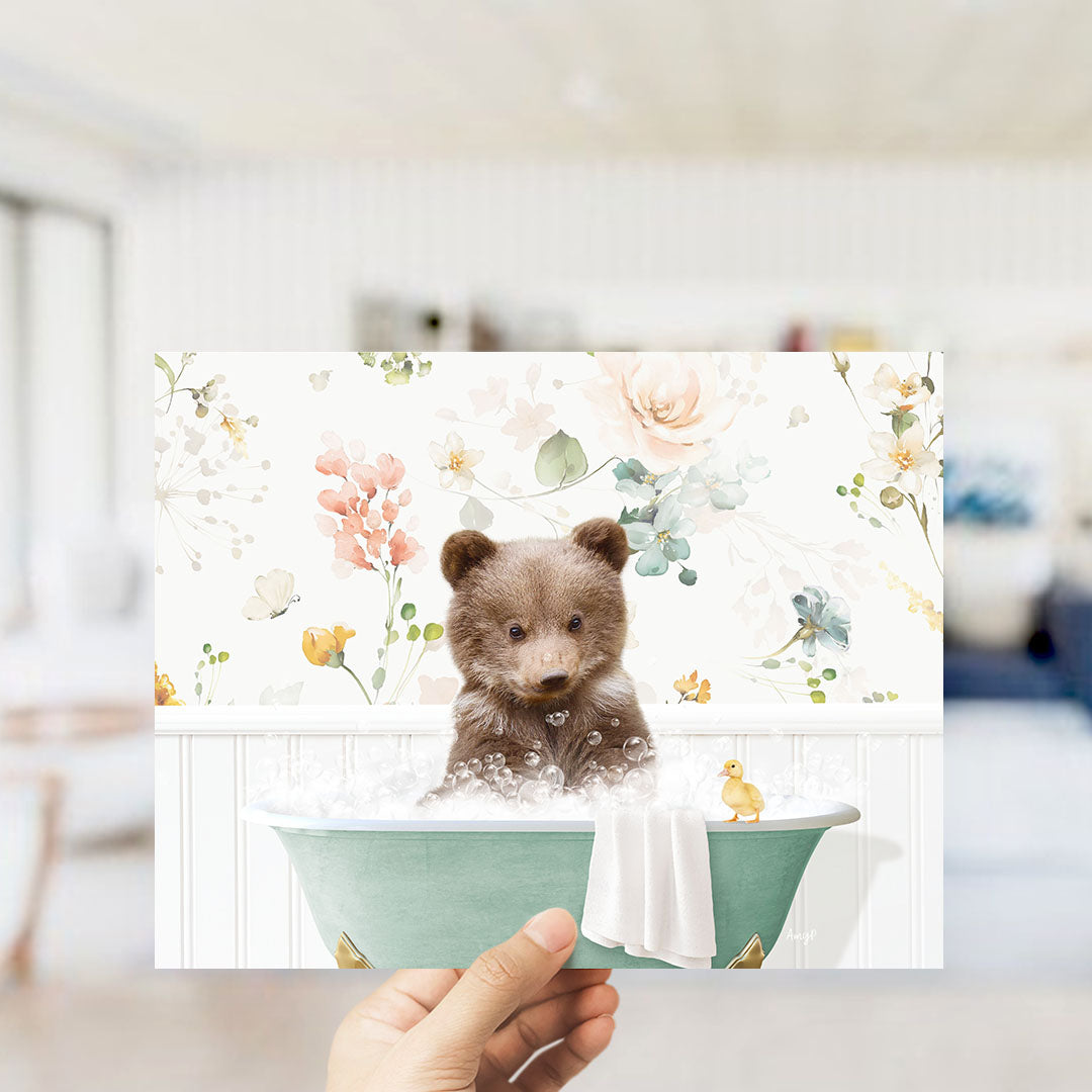 A hand holding a framed print featuring a cute brown bear sitting in a bathtub filled with bubbles, surrounded by a floral pattern.