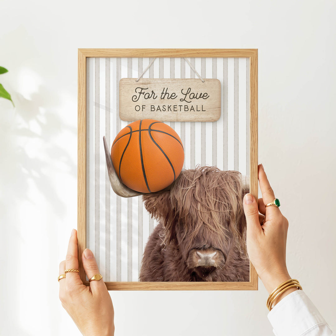 A person's hands are holding a framed picture of a Highland cow with a basketball on its head.