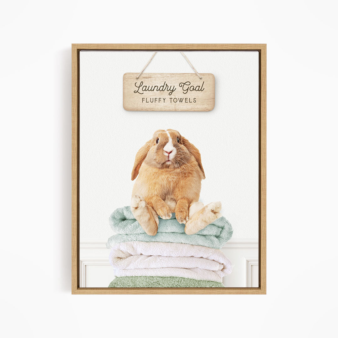 A wooden sign with the text "Laundry Goal Fluffy Towels" hangs above a stack of folded towels, with a stuffed rabbit sitting on top of the towels.