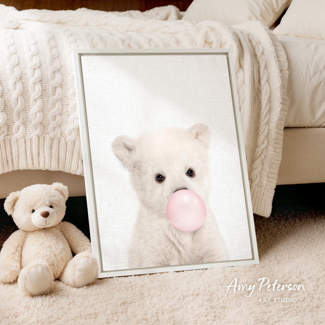 A framed image of a white polar bear blowing a bubble with a pink bubble gum bubble is displayed on a bed, accompanied by a teddy bear.