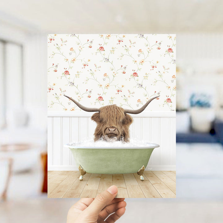 A hand holds up a framed image depicting a Highland cow with long horns sitting in a green bathtub filled with water.