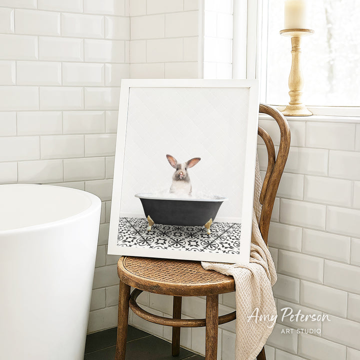 A framed image of a rabbit sitting in a bathtub, surrounded by a patterned tile floor, is displayed on a wooden chair in a bathroom setting.
