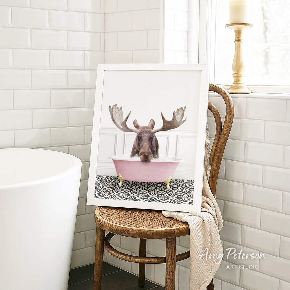 A framed image of a moose sitting in a pink bathtub, with a towel draped over the back of a wooden chair in a bathroom setting.