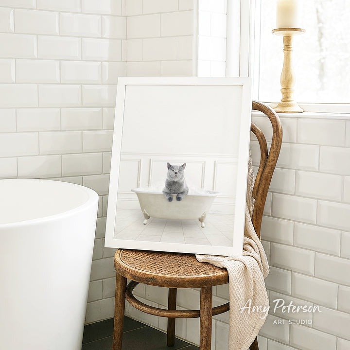 A framed image of a gray cat sitting in a white bathtub, with a wooden chair and a white tiled wall in the background.