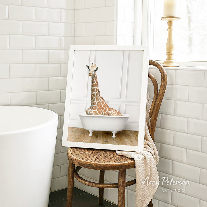 A framed image of a giraffe sitting in a bathtub is displayed on a wooden chair in a bathroom setting.