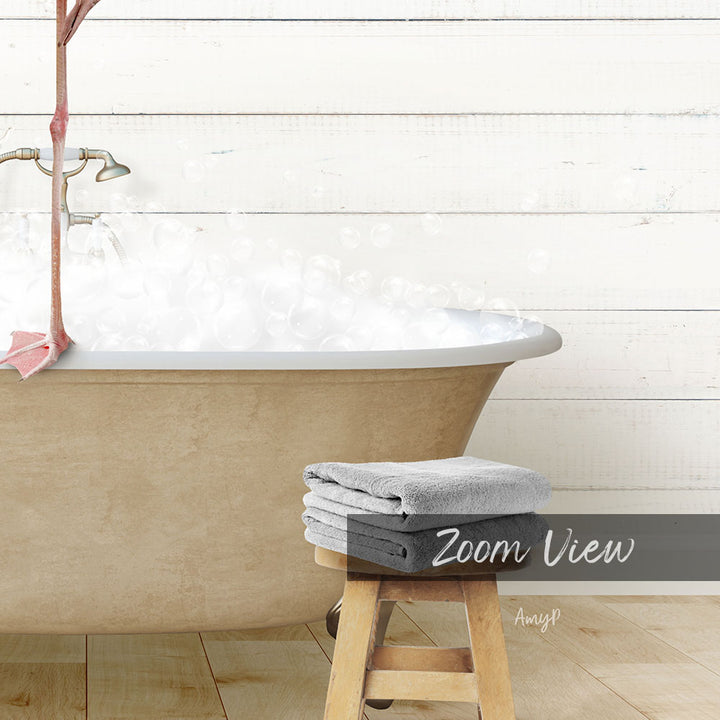A bathtub filled with bubbles, a wooden stool, and a stack of towels, all set against a white wooden wall.