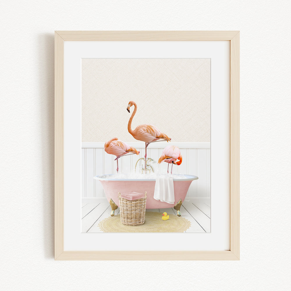 A framed image depicting three pink flamingos standing in a pink bathtub, with a basket of bath toys and a yellow rubber duck nearby.