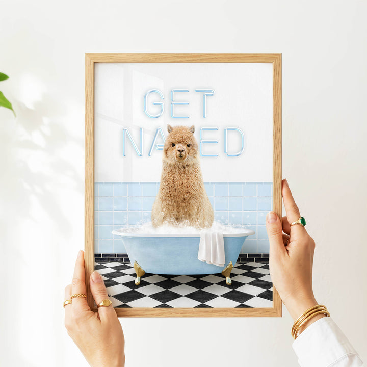 A person is holding up a framed image of a brown alpaca sitting in a blue bathtub, with the words "GET NAKED" written on the frame.