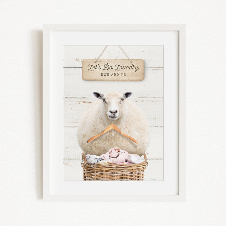 A framed image of a sheep sitting in a basket filled with laundry, with a sign above it that says "Let's Do Laundry EWS AND ME".