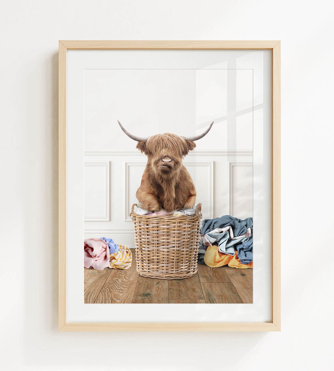 A Highland cow with long horns is sitting in a wicker basket filled with clothes, placed on a wooden floor.